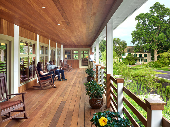 A large porch with Garapa wood decking, soffits, railings and vertical siding. The porch has rocking chairs and large windows.