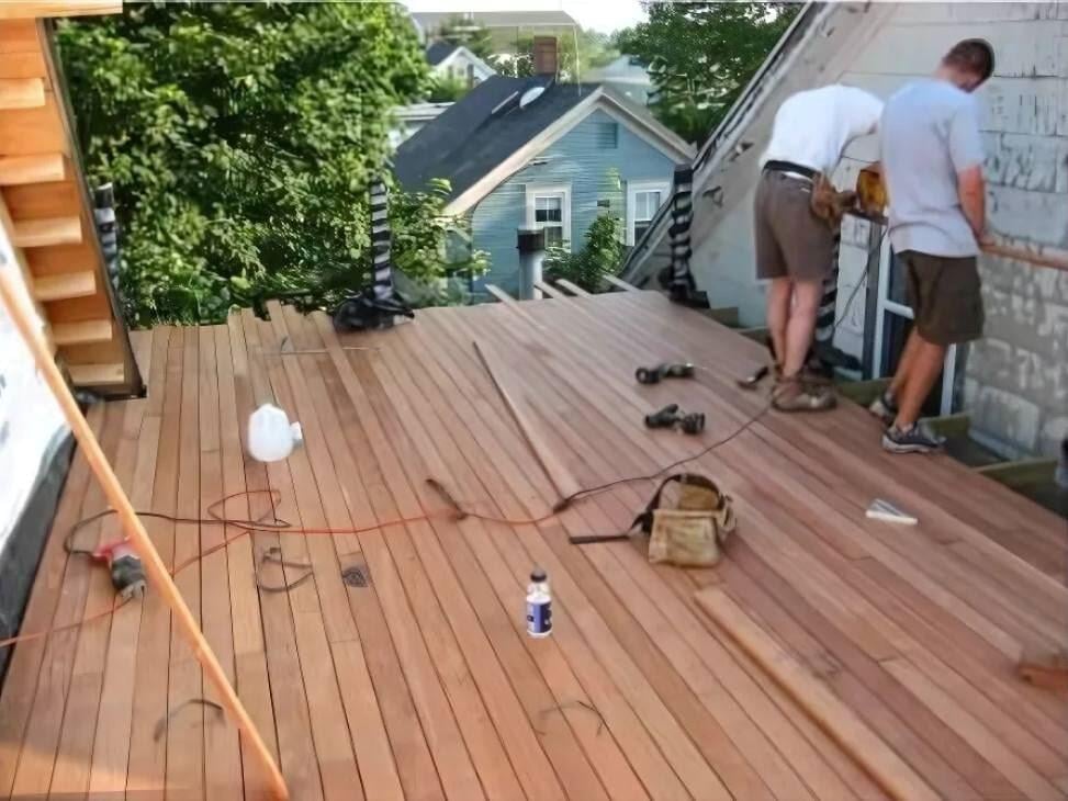 Mataverde Ipe wood small rooftop deck under construction with two workers and tools, with a view of the rooftops of houses and trees in the background