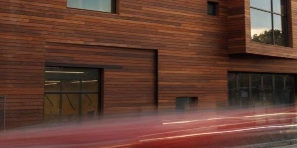 A close up of the street facing exterior of a contemporary building with Ipe wood rainscreen siding. The building has large windows and a few small, square openings. There is a street in front of the building with a blurred image of a car passing by.