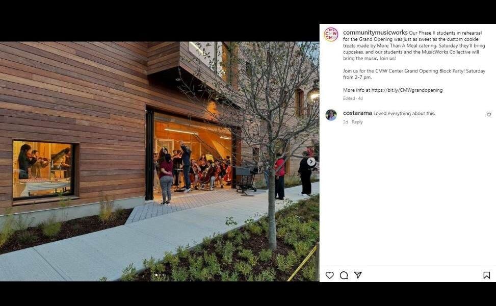 A group of musicians rehearse inside the music school with Ipe siding surrounding the with large windows and a large, open glass doorway. People are on the sidewalk in the doorway listening to the performance. ing, There are trees and greenery planted in the foreground along the sidewalk. IG @CommunityMusicWorks courtesy Sebastian Bach