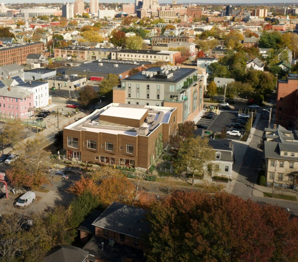 A rendering of an aerial view of Community MusicWorks,  a modern building with a flat roof and a wood-paneled facade, Located in a densely populated urban area with many other buildings and trees. The surrounding trees are showing vibrant autumn colors.